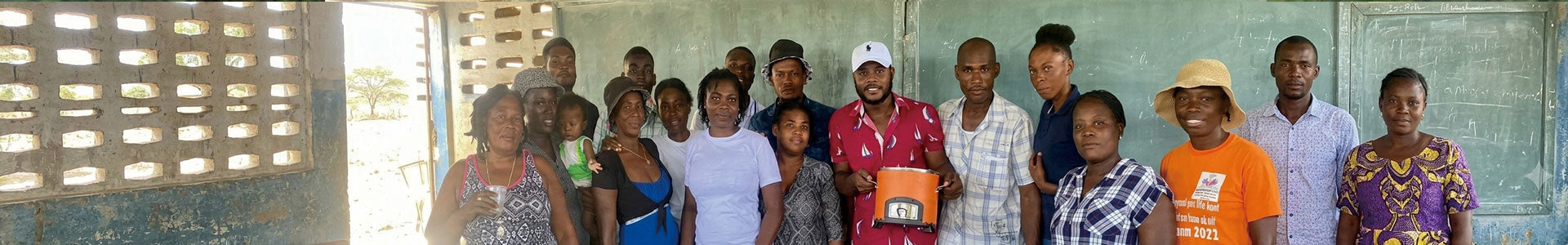 A group of people stand together for a photograph inside a classroom with a blackboard. A man in the center holds an orange charcoal stove.