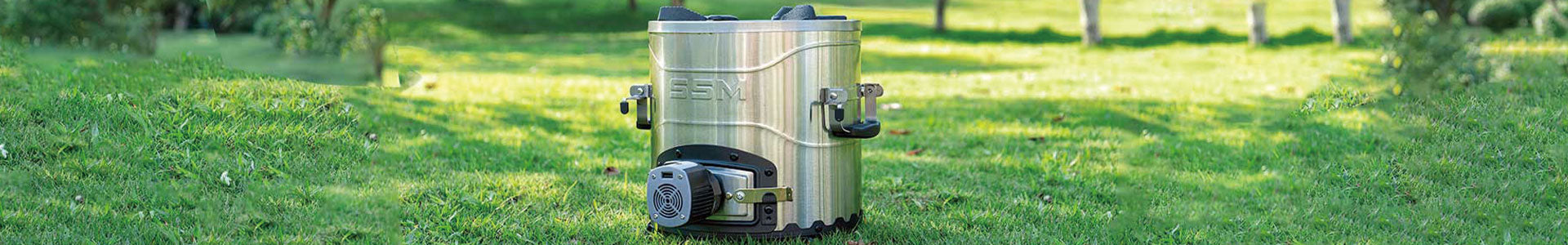 An SSM stainless steel pellet stove equipped with high-efficiency pellet-burning technology, set outdoors on a vibrant, sunlit green lawn against a soft-focus background of trees.