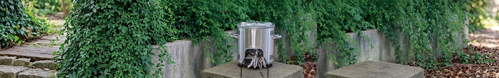 An SSM stainless steel wood-burning stove, featuring the efficient and clean-burning technology showcased in 'wood-stove.jpg', is set upon a concrete pedestal in a lush garden, surrounded by climbing greenery and concrete planters.