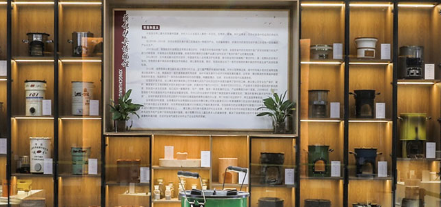 A panoramic view of the SSM product showroom and corporate exhibition hall, featuring illuminated display shelves showcasing a diverse collection of historical and modern biomass cookstoves, components, and company history panels against an exposed brick and grey wall backdrop.