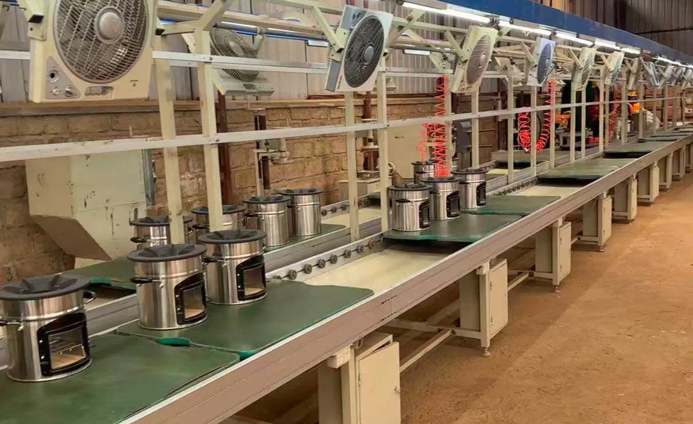 A clean, modern production line in SSM's local assembly plant in Rwanda, featuring assembled stainless steel biomass cookstoves arranged sequentially along a long conveyor belt system equipped with overhead cooling fans.