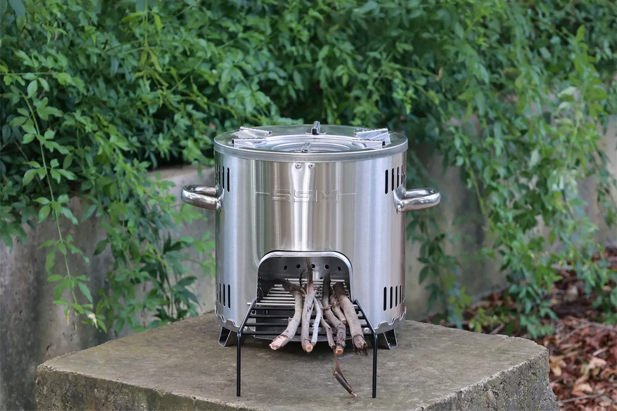 An SSM stainless steel wood stove placed on a stone path in a forest setting for outdoor camping.