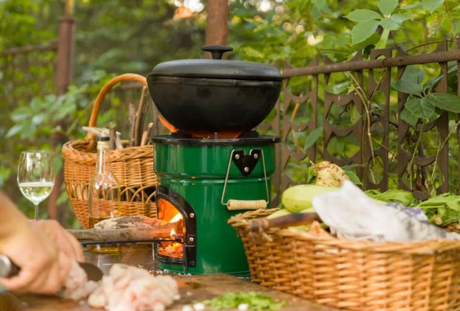 An outdoor scene featuring a green SSM wood burning stove in use with a heavy pot on top, set up on a wooden table alongside baskets, a wine glass, and food preparation elements.