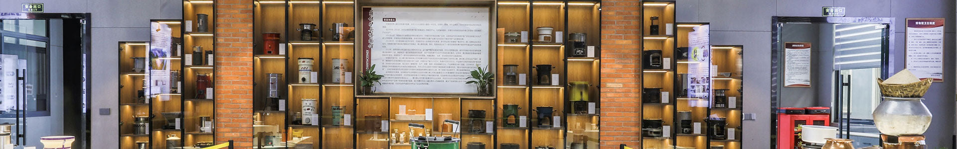 A panoramic view of the SSM product showroom and corporate exhibition hall, featuring illuminated display shelves showcasing a diverse collection of historical and modern biomass cookstoves, components, and company history panels against an exposed brick and grey wall backdrop.